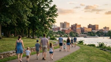 Families walk a tree-lined riverfront path at golden hour with a city skyline across the water (illustrative)