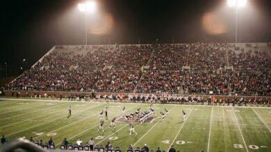A college football night game viewed from the upper stands, floodlights over the field and a filled grandstand behind (illustrative)
