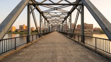 Looking down the deck of an early-1900s steel truss pedestrian bridge over a river toward a downtown skyline at golden hour (illustrative)